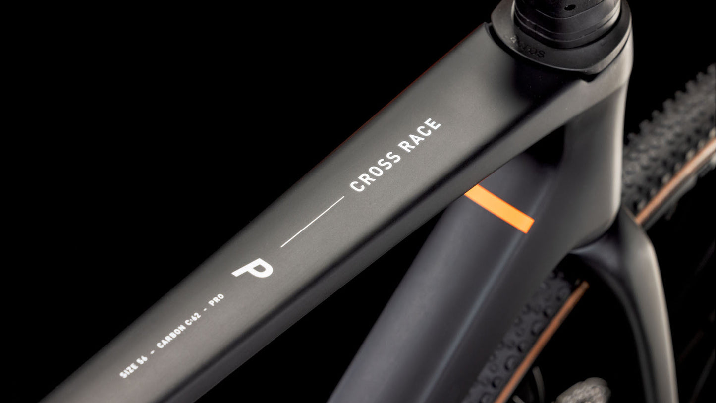 Nahaufnahme eines mattschwarzen CUBE Cross Race C:62 Pro carbon'n'orange 2025 Rahmens, "Cross Race" und Größe M in weiß auf dem Oberrohr, dezenter oranger Akzent, Schotterreifen im Hintergrund, ausgestattet mit Shimano 105 Di2.
