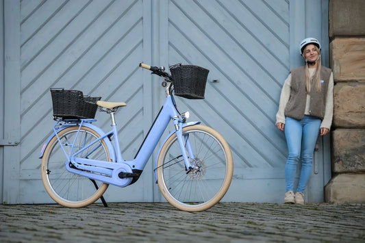 Hellblaues CUBE City-Bike mit Körben an einer blauen Holztür, daneben eine Frau mit Helm