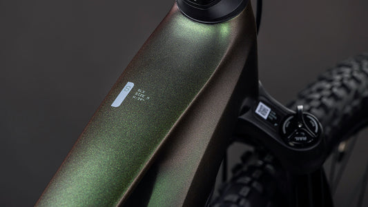 Nahaufnahme des metallisch glänzenden, dunkelgrünen Rahmens des CUBE Stereo Hybrid ONE22 SLX 800 stellar'n'green 2026 E-Fully, die ein kleines Etikett und einen Teil des Lenkers vor einem dunklen, verschwommenen Hintergrund zeigt.
