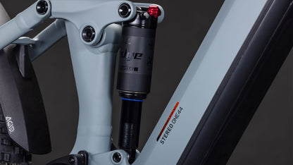 Nahaufnahme der Hinterradfederung des CUBE Stereo Hybrid ONE44 Pro 800. Zu sehen sind der Stoßdämpfer, der Rahmen mit der Aufschrift "STEREO ONE44" und die Shimano XT-Schaltung in Hellgrau und Orange vor dunklem Hintergrund.