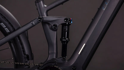 Nahaufnahme des CUBE Stereo Hybrid ONE44 EXC 800 FE irongrey'n'chrome 2026 Rahmens, mit Fokus auf die Hinterradaufhängung und den Stoßdämpfer, wobei ein Teil des Hinterrads und der Kette vor einem dunklen Hintergrund sichtbar ist.