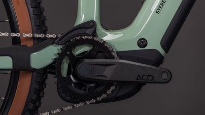 Nahaufnahme der High-Performance-Carbon-Kurbelgarnitur und des schwarzen ACID-Kurbelarms des CUBE Stereo Hybrid ONE44 HPC Race 800 driedherbs'n'black 2026, die am mintgrünen Rahmen befestigt sind, wobei ein Teil des Hinterrads und des Reifens sichtbar ist.