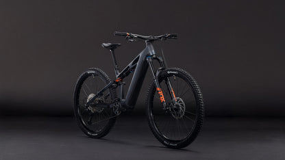 Das CUBE Stereo Hybrid ONE44 HPC SLX 800 slabgrey'n'orange 2026 Mountainbike mit Bosch CX Motor, Shimano XT Schaltung, Scheibenbremsen und orangen Akzenten steht aufrecht und schräg links vor einem dunklen Hintergrund.