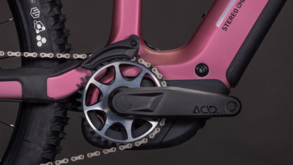 Nahaufnahme der CUBE Stereo Hybrid ONE44 HPC SLX 800 shiftblush'n'art 2026 Kurbelgarnitur, des ACID-gekennzeichneten Pedalarms und des Kettenblatts, wobei ein Teil des Hinterradreifens und des Rahmenlogos zu sehen ist, was die fortschrittliche Integration des Bosch CX-Motors zeigt.