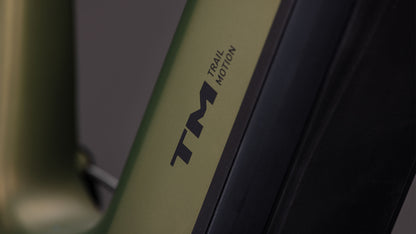 Nahaufnahme eines CUBE Stereo Hybrid ONE44 HPC TM 800 reedgreen'n'matrix 2026 E-Mountainbike mit mattgrünem und schwarzem Rahmen, auf dem der Text TM TRAIL MOTION in schwarzer Schrift zu lesen ist. Der Hintergrund ist unscharf und grau.