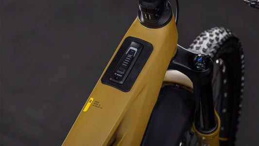 Nahaufnahme des Bedienfelds am Oberrohr des CUBE Stereo Hybrid ONE77 HPC Race 800 willowgreen'n'black 2026 E-Mountainbikes. Zu sehen sind Tasten, eine LED-Anzeige, Teile des Lenkers und der Hinterreifen.