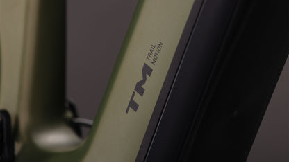 Nahaufnahme des CUBE Stereo Hybrid ONE77 HPC TM 800 reedgreen'n'matrix 2026 Rahmens mit "TM" und "Trail Motion" in schwarz, neben einer schwarzen Sram GX Eagle Getriebekomponente. Der Hintergrund ist unscharf.