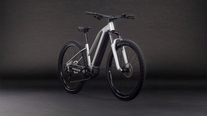 Das CUBE Reaction Hybrid ONE 800 sleekgrey'n'prism Trapeze 2026 Elektrofahrrad mit mattsilbernem Rahmen, Bosch-Motor, dicken Reifen und geradem Lenker wird vor einem dunklen Hintergrund mit Farbverlauf gezeigt.
