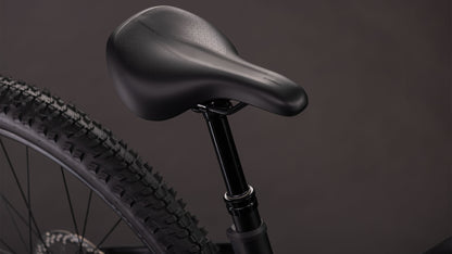 Nahaufnahme des schwarzen Sattels des CUBE Cube Reaction Hybrid Pro 800 blackline Trapeze 2026 auf der Sattelstütze, wobei ein Teil des Hinterrads und des Reifens des E-Mountainbikes vor einem dunklen Hintergrund zu sehen ist.