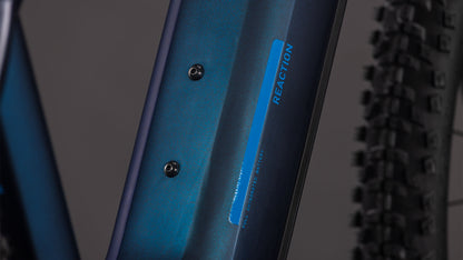 Nahaufnahme des Cube Reaction Hybrid Pro 800 nebular'n'dazzle 2026 Rahmens von CUBE mit zwei Befestigungsschrauben und REACTION Schriftzug in Blau. Im Hintergrund ist eine knorrige Fahrradreifenkante zu sehen.