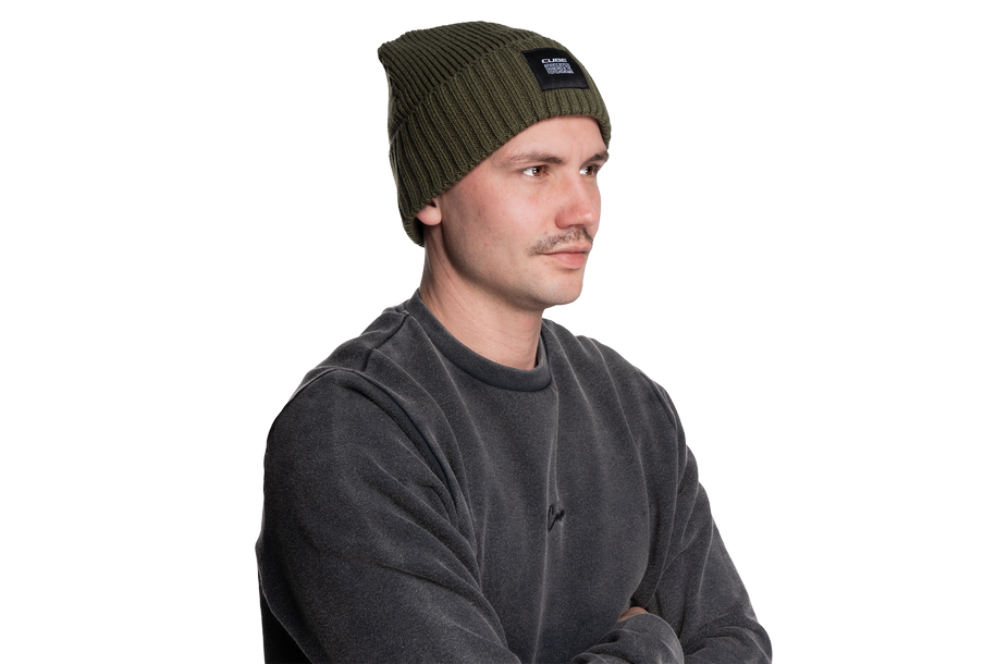 Ein Mann mit Cube CUBE Beanie oliv und dunkelgrauem Sweatshirt steht mit verschränkten Armen und Blick zur Seite vor einem weißen Hintergrund und verkörpert den coolen, modernen Stil Göttingens.