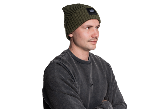 Ein Mann mit Cube CUBE Beanie oliv und dunkelgrauem Sweatshirt steht mit verschränkten Armen und Blick zur Seite vor einem weißen Hintergrund und verkörpert den coolen, modernen Stil Göttingens.