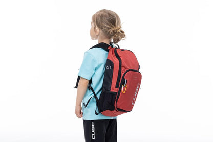 Ein junges Kind mit blonden Haaren in einem Dutt, einem hellblauen Hemd und einer schwarzen Hose, steht seitlich mit dem CUBE Rucksack PURE 6 ROOKIE red'n'black von CUBE vor einem schlichten weißen Hintergrund.