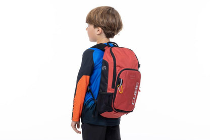 Ein Junge mit kurzen braunen Haaren trägt ein blau-orangefarbenes Hemd und hat einen CUBE Rucksack PURE 6 ROOKIE red'n'black von CUBE bei sich, der vor einem schlichten weißen Hintergrund steht.