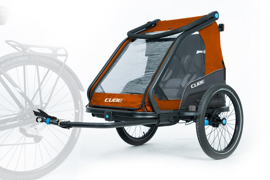 Der Cube CUBE Kinder Fahrradanhänger Double CMPT X Actionteam, in orange und schwarz, verfügt über zwei große Räder, einen durchsichtigen vorderen Insektenschutz und Sitzplätze im Inneren. Er wird links an einem teilweise sichtbaren Fahrrad befestigt gezeigt.
