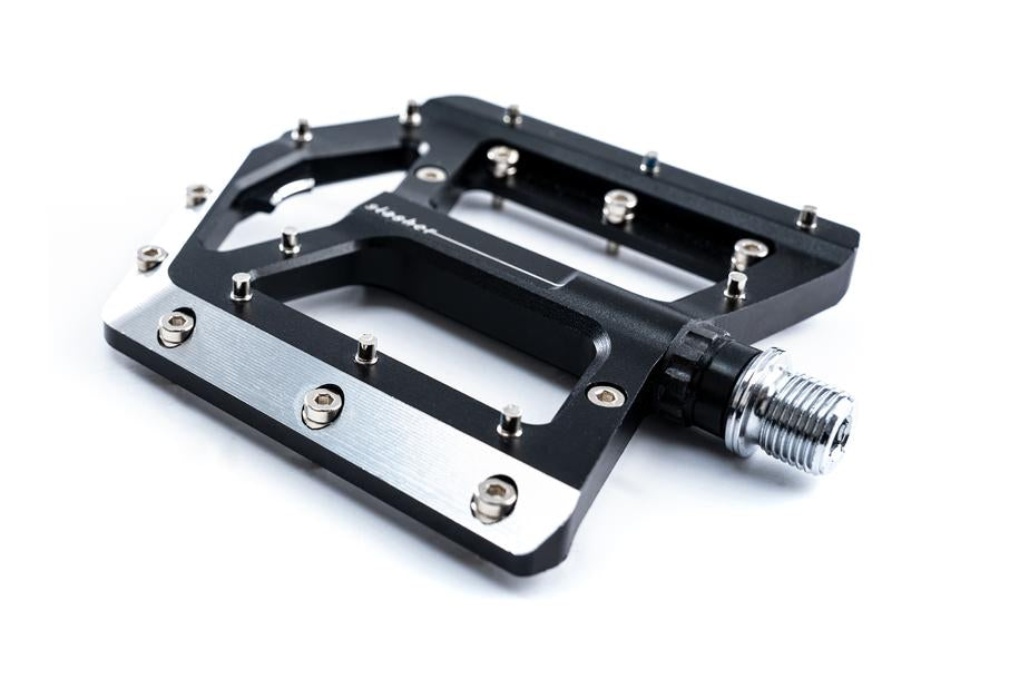 Eine Nahaufnahme des Cube CUBE Pedale SLASHER Flatpedals mit großer schwarz-silberner Plattform und verstellbaren Pins für optimalen Halt, fotografiert vor weißem Hintergrund.