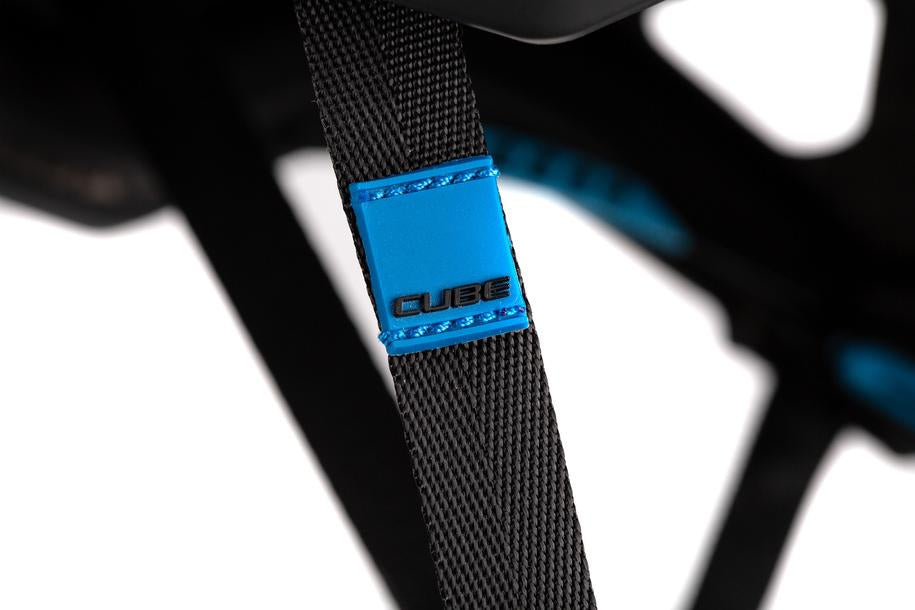Nahaufnahme eines schwarzen verstellbaren Riemens mit blauem Akzent und der Aufschrift Cube vom CUBE Helm BADGER / MTB Enduro Helm. Der unscharfe Hintergrund hebt die Nähte des Armbands und das eindeutige MTB Enduro Helm Branding hervor.