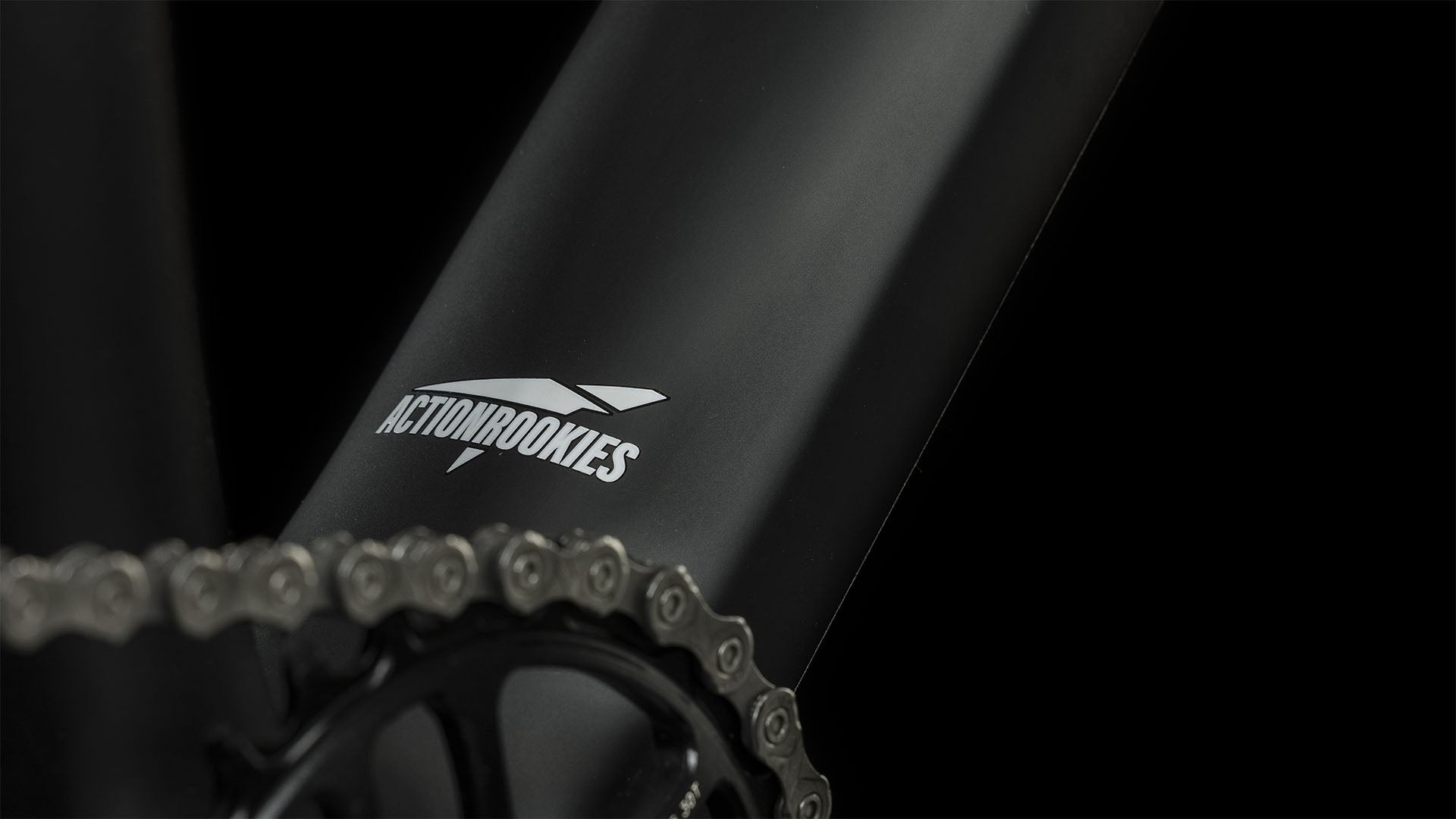 Nahaufnahme des mattschwarzen CUBE Elite 240 C:62 Pro carbon'n'blue'n'red 2024-Rahmens mit weißem CUBE-Logo in der Nähe der Fahrradkette und des Kettenblatts vor einem schwarzen Hintergrund.