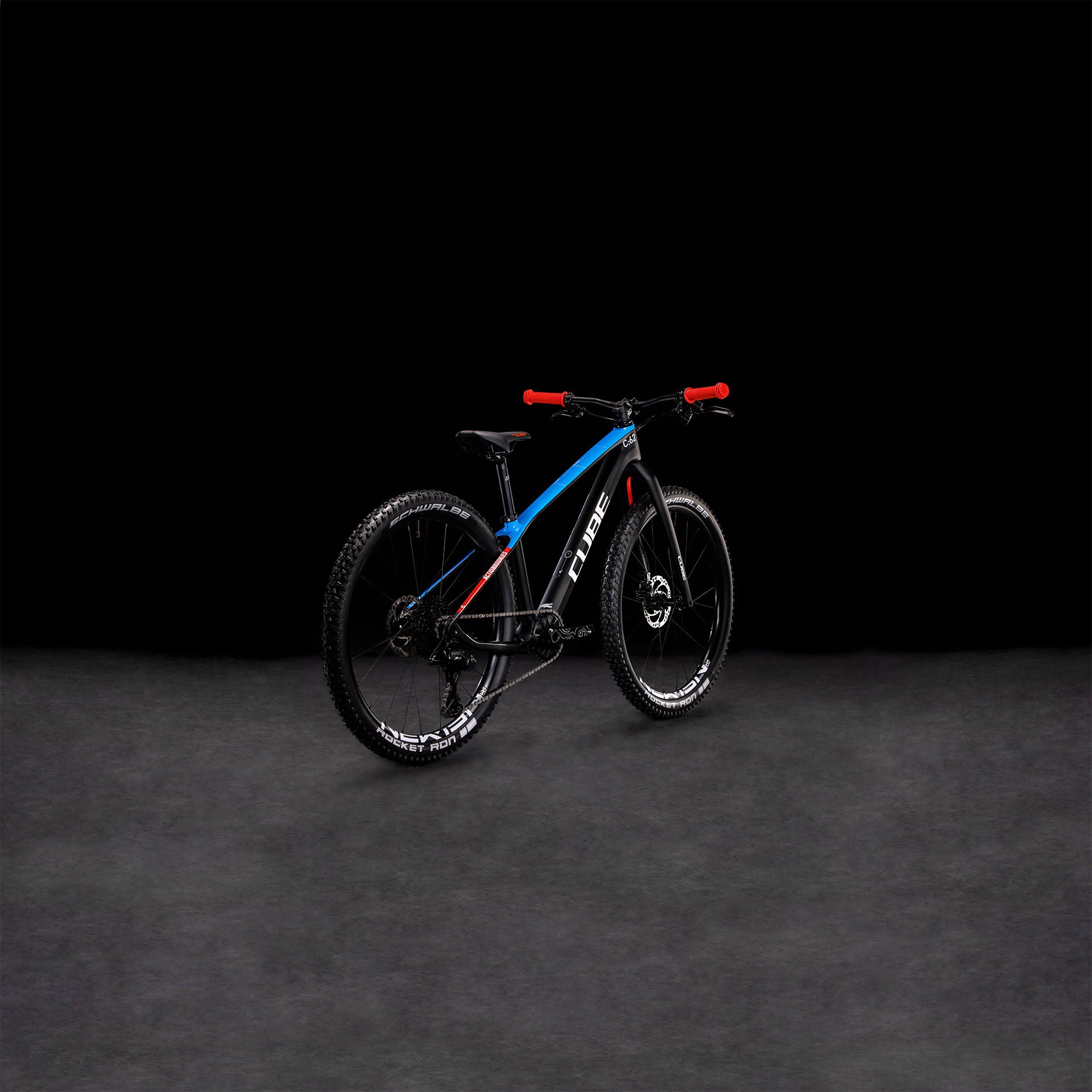 Das CUBE Elite 240 C:62 Pro carbon'n'blue'n'red 2024, ein CUBE Race-Mountainbike mit Vollcarbonrahmen in Blau und Schwarz, rotem Lenker und dicken Reifen, steht auf einer dunklen Fläche vor schwarzem Hintergrund.