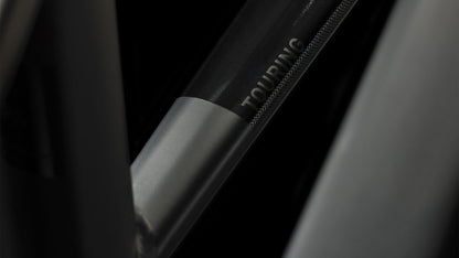 Nahaufnahme eines CUBE Touring Hybrid Pro 625 black'n'metal Trapeze 2024 Rahmenrohrs mit dem Aufdruck "TOURING", der ein elegantes dunkles Metallfinish mit subtilen Licht- und Schatteneffekten zeigt.