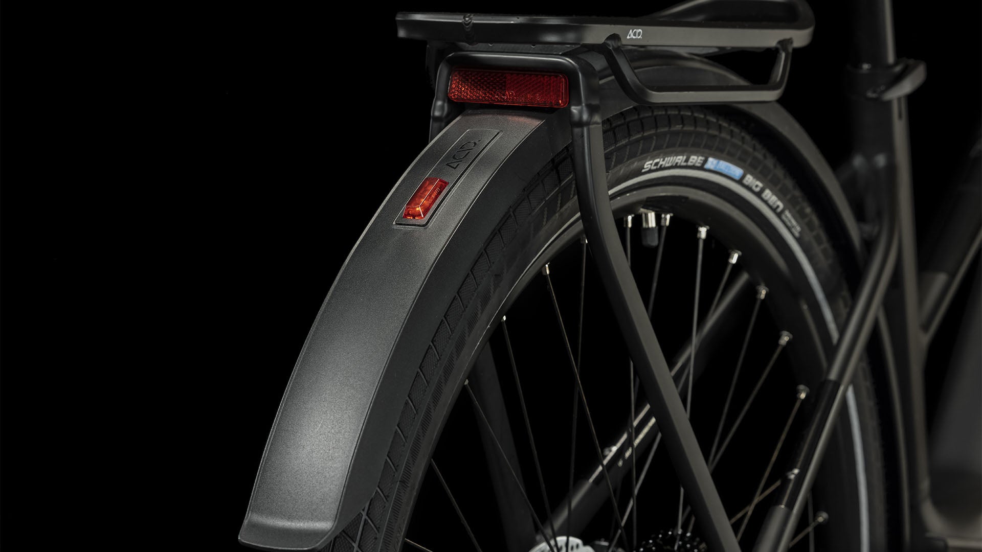 Nahaufnahme des Hinterrads eines CUBE Touring Hybrid Pro 625 black'n'metal Trapeze 2024, wobei das Schutzblech, der Reifen, die Speichen, der Gepäckträger und der rote Reflektor vor einem schwarzen Hintergrund hervorgehoben werden.