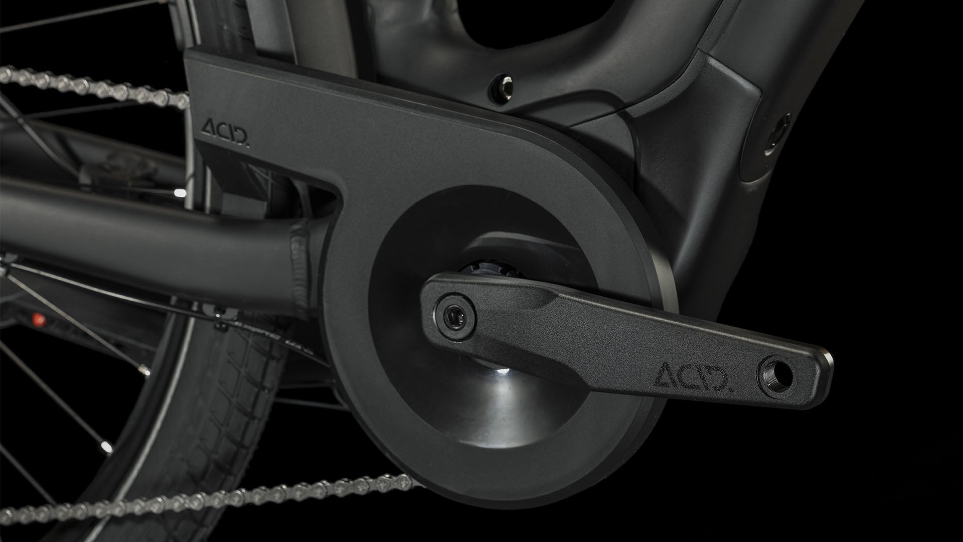 Nahaufnahme des schwarzen Kurbelarms und Kettenschutzes des CUBE Touring Hybrid Pro 625 black'n'metal Trapeze 2024 mit ACID-Logo, Bosch Performance Motor und Shimano 11-Gang Schaltung vor einem dunklen Hintergrund, der das Hinterrad und die Kette hervorhebt.