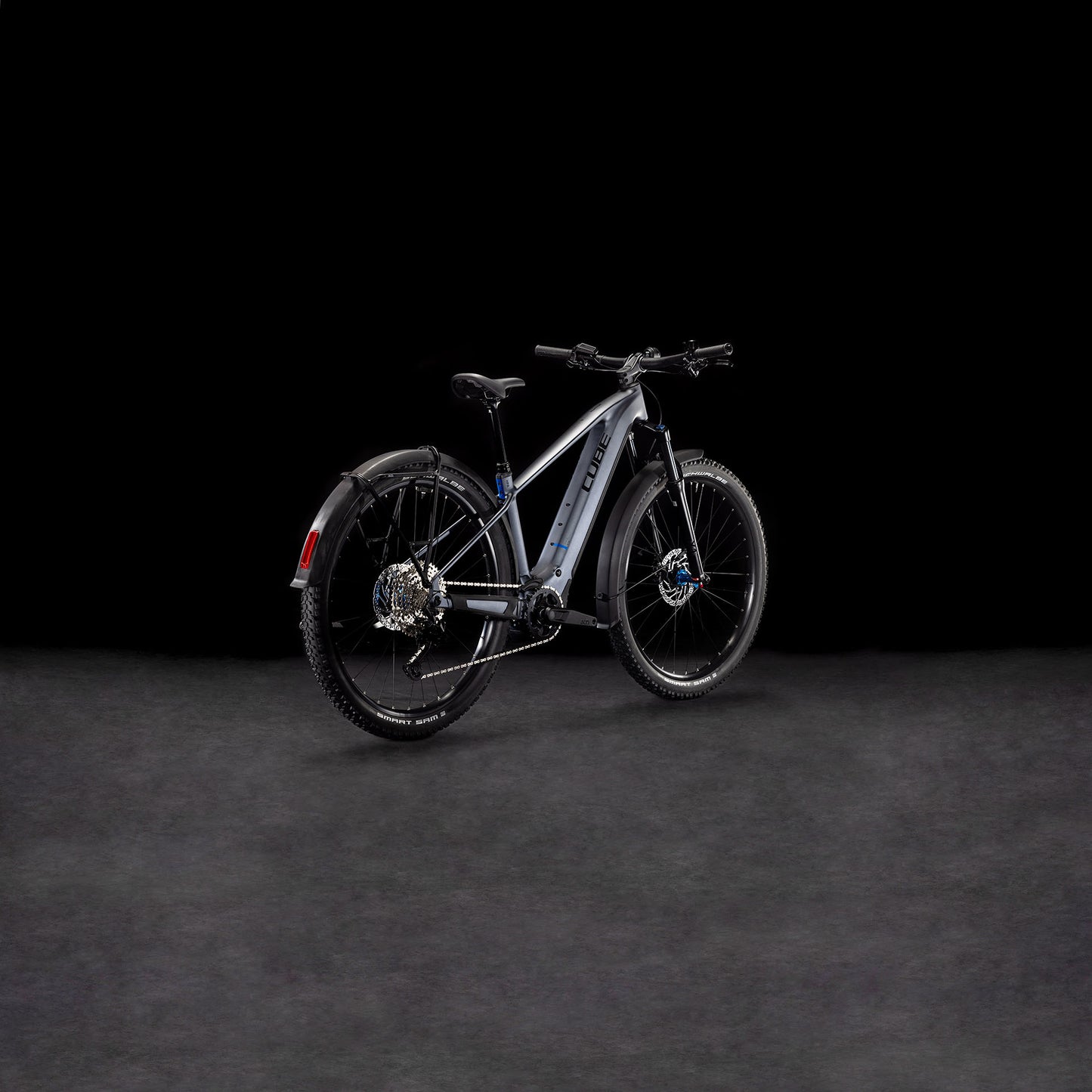 Das CUBE Reaction Hybrid Pro 800 Allroad metallicgrey'n'black 2025, ein modernes E-Bike mit dicken Reifen, Schutzblechen und hydraulischen Scheibenbremsen, wird von hinten links auf dunklem Hintergrund gezeigt.