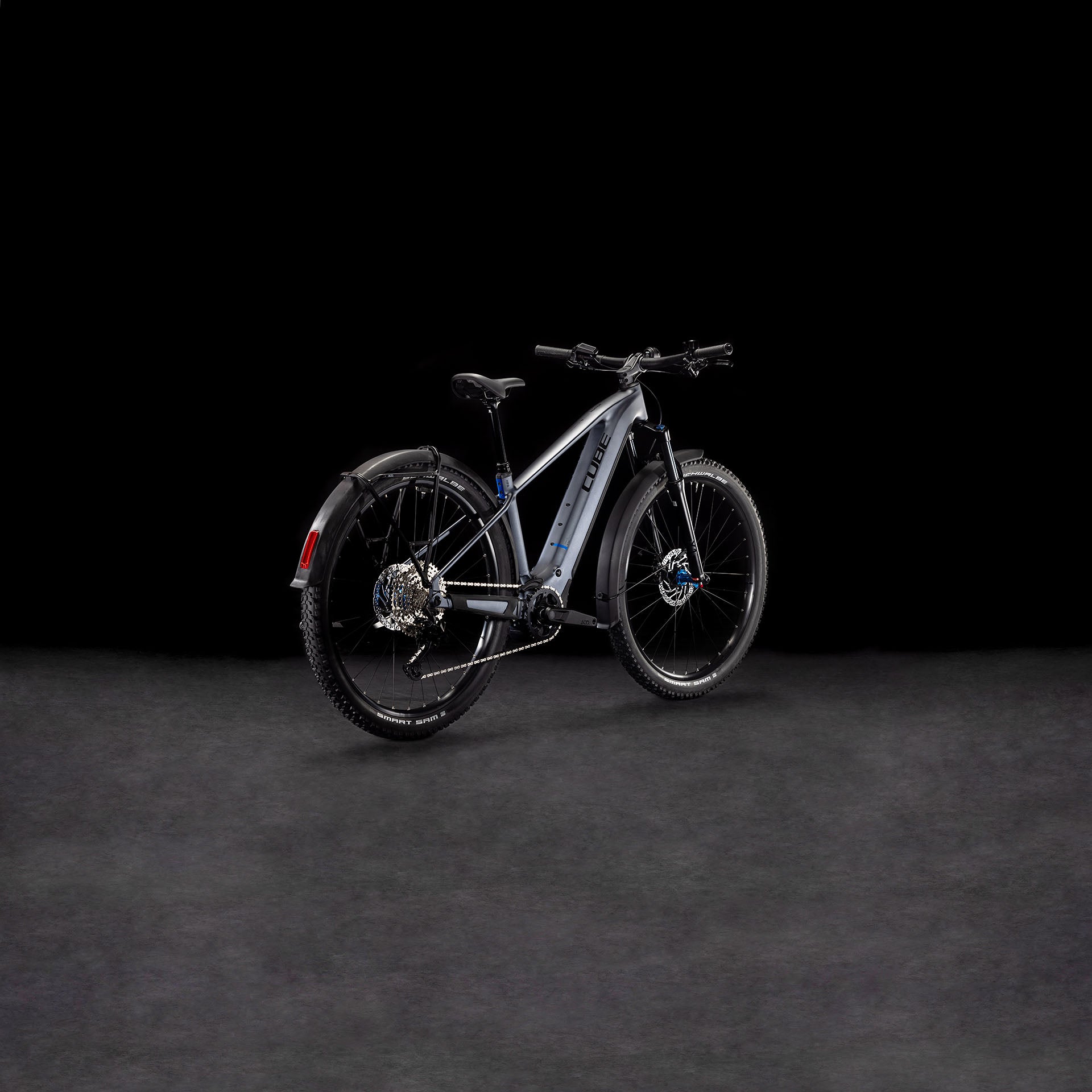 Das CUBE Reaction Hybrid Pro 800 Allroad metallicgrey'n'black 2025, ein modernes E-Bike mit dicken Reifen, Schutzblechen und hydraulischen Scheibenbremsen, wird von hinten links auf dunklem Hintergrund gezeigt.