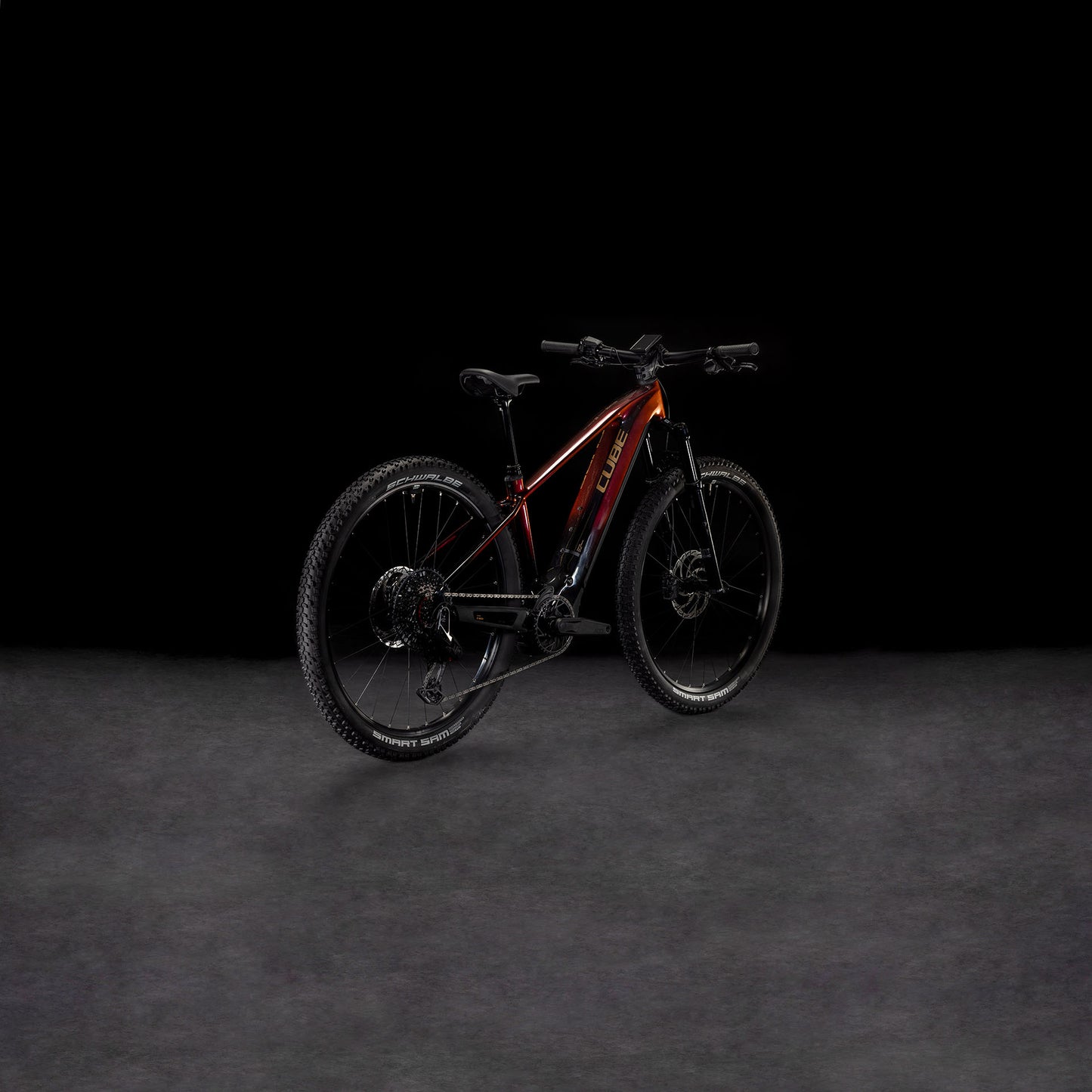 Das CUBE Reaction Hybrid SLT 800 solareclipse'n'bronze 2025 mit dicken Reifen und einem Bosch CX-Motor wird vor einem schattigen Hintergrund gezeigt, wobei der rot-schwarze Rahmen leicht nach rechts gedreht ist, um die Seiten- und Hinterräder zu präsentieren.