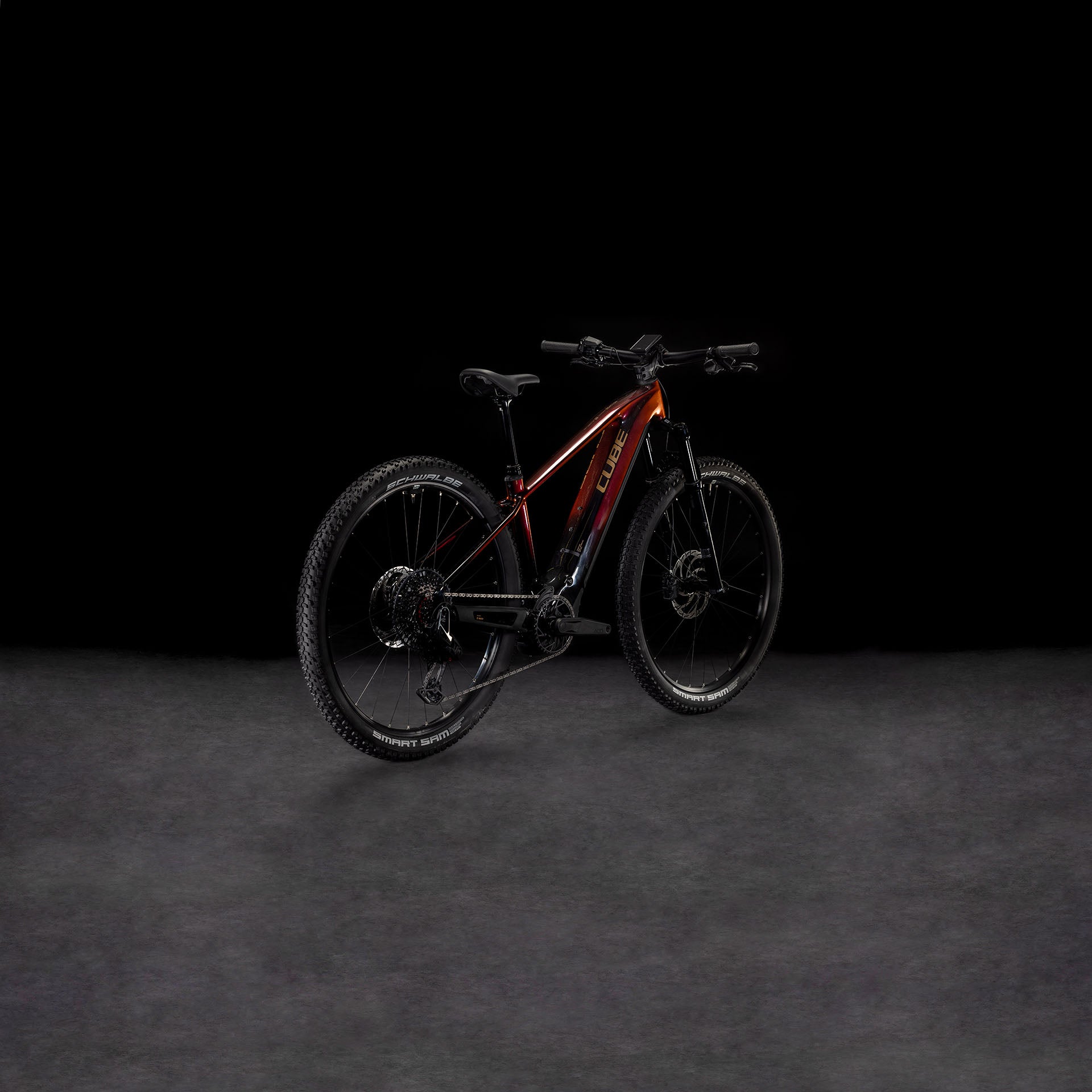 Das CUBE Reaction Hybrid SLT 800 solareclipse'n'bronze 2025 mit dicken Reifen und einem Bosch CX-Motor wird vor einem schattigen Hintergrund gezeigt, wobei der rot-schwarze Rahmen leicht nach rechts gedreht ist, um die Seiten- und Hinterräder zu präsentieren.