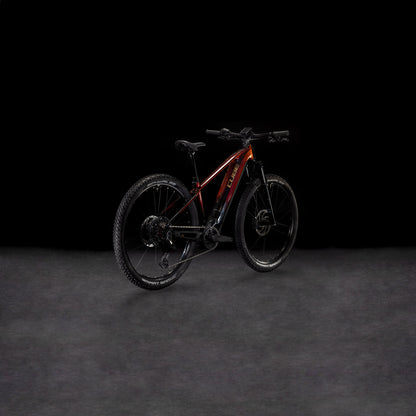 Das CUBE Reaction Hybrid SLT 800 solareclipse'n'bronze 2025 mit dicken Reifen und einem Bosch CX-Motor wird vor einem schattigen Hintergrund gezeigt, wobei der rot-schwarze Rahmen leicht nach rechts gedreht ist, um die Seiten- und Hinterräder zu präsentieren.