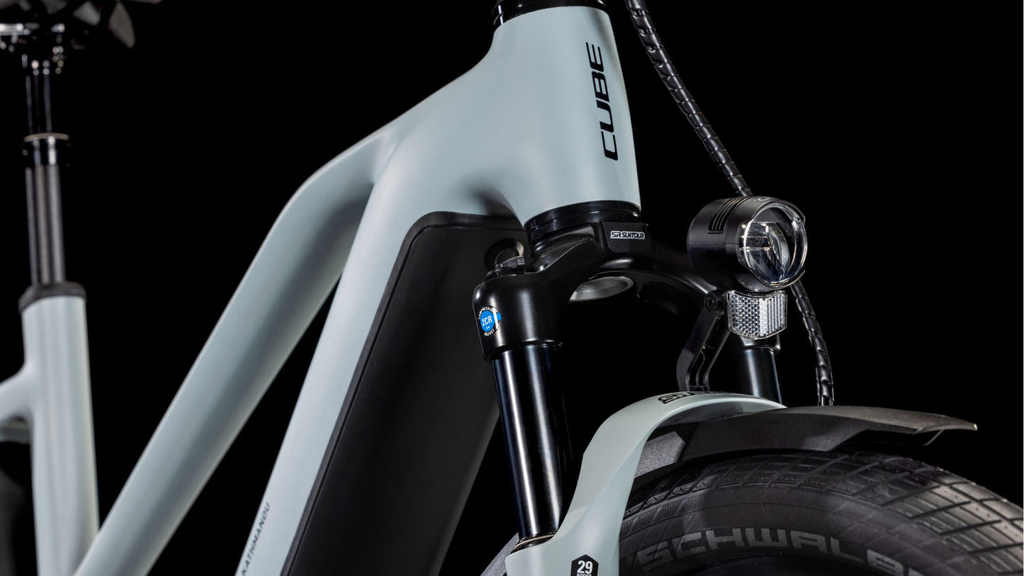 Die Nahaufnahme des CUBE Kathmandu Hybrid EX 800 haze´n´black Trapeze 2025 zeigt die Federgabel, den Scheinwerfer, den dicken Reifen und den Rahmen mit CUBE-Branding auf mattem hellgrauem Hintergrund. Ausgestattet mit einem Bosch CX Motor für kraftvolle Leistung.