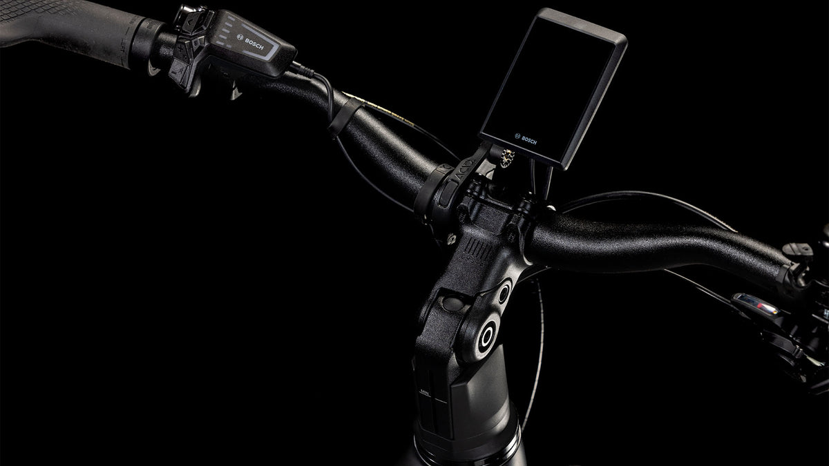 Nahaufnahme des CUBE Kathmandu Hybrid EXC 800 black'n'prism Trapeze 2025 Lenkers mit Bosch-Digitalanzeige, Bedientasten, strukturiertem Griff und Bremshebel vor schwarzem Hintergrund.