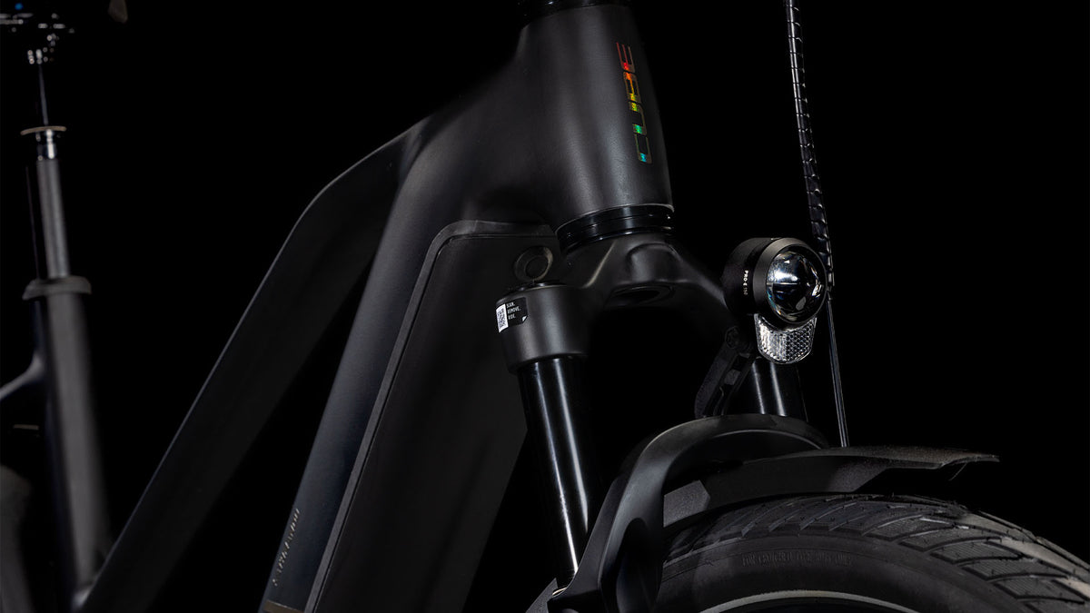 Die Nahaufnahme des CUBE Kathmandu Hybrid EXC 800 black'n'prism Trapeze 2025 zeigt die mattschwarze Front mit Federgabel, Scheinwerfer und einem Teil des Vorderreifens vor einem schwarzen Hintergrund.