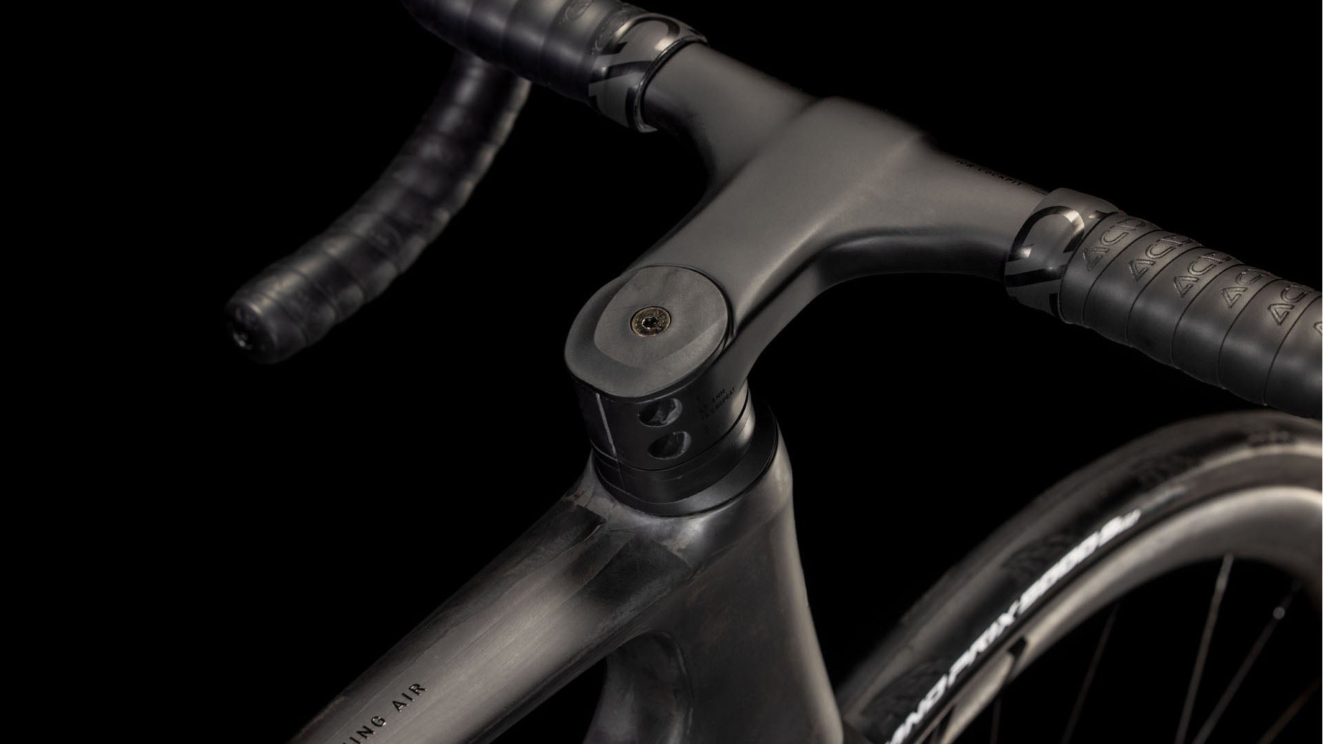 Nahaufnahme des Cube Litening AIR C:68X Race blackline 2025 von CUBE, die den aerodynamischen Lenker, den Vorbau und das Vorderrad vor einem schwarzen Hintergrund zeigt. Der Carbon-Rahmen sieht mit dem schwarzen, mit Tape umwickelten Lenker schlank und modern aus.