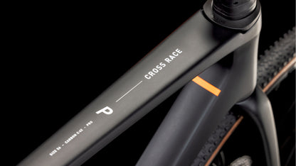 Nahaufnahme eines mattschwarzen CUBE Cross Race C:62 Pro carbon'n'orange 2025 Rahmens, "Cross Race" und Größe M in weiß auf dem Oberrohr, dezenter oranger Akzent, Schotterreifen im Hintergrund, ausgestattet mit Shimano 105 Di2.