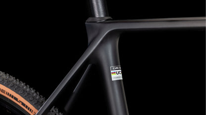 Nahaufnahme des mattschwarzen Cube Cross Race C:62 Pro carbon'n'orange 2025 Carbon-Rahmens von CUBE. Zu sehen sind der Übergang zwischen Sitz- und Oberrohr, ein UCI-geprüfter Aufkleber und ein Noppenreifen. Shimano 105 Di2 kompatibel.
