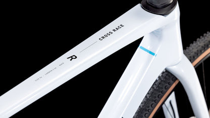 Nahaufnahme eines CUBE Cube Cross Race C:62 Race polarwhite'n'blue 2025 Rahmens mit der Aufschrift CROSS RACE, der klare Linien, eine glatte Oberfläche und einen Noppenreifen vor einem schwarzen Hintergrund zeigt.