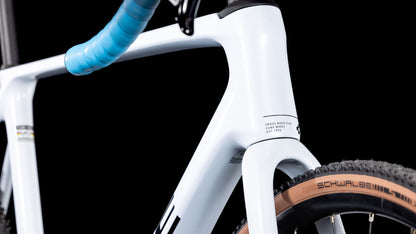 Nahaufnahme des CUBE Cross Race C:62 Race polarwhite'n'blue 2025 Rahmens mit blauem Lenker, braunen Schwalbe Reifen und Sram Rival AXS, vor schwarzem Hintergrund.