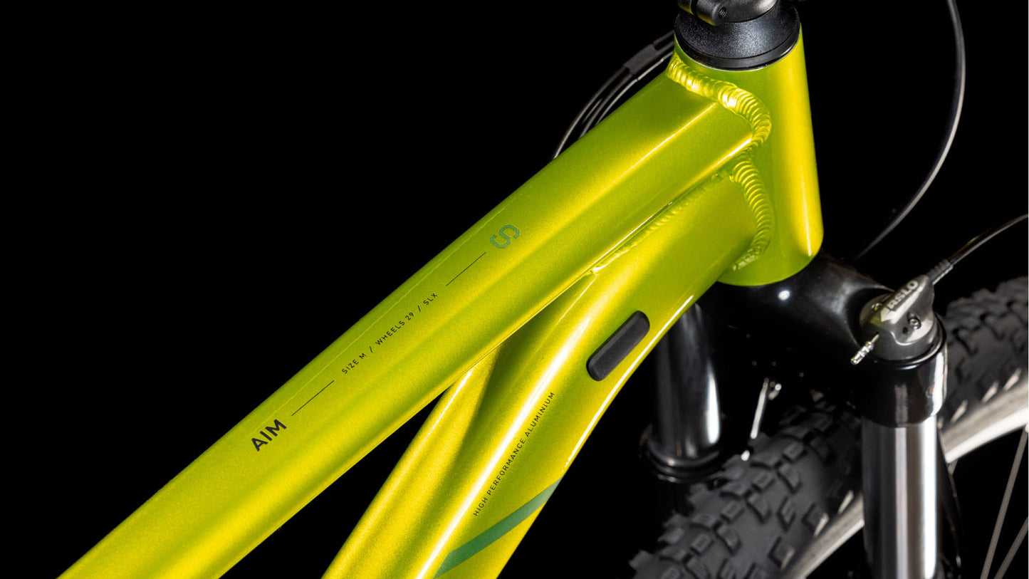 Nahaufnahme des CUBE Aim SLX Flashlime´n´Juniper 2025 Mountainbike-Rahmens, AIM-Modellbezeichnung, Vorderradgabel, Teil des Reifens und seiner hydraulischen Shimano-Scheibenbremsen vor schwarzem Hintergrund.