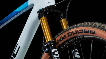 Nahaufnahme des weißen Rahmens mit blauen Akzenten des CUBE Phenix C:68X SLT teamline 2025, der Fox Float 32 Factory SC-Gabel und des Racing Ray-Reifens mit hellbraunen Flanken vor einem schwarzen Hintergrund.