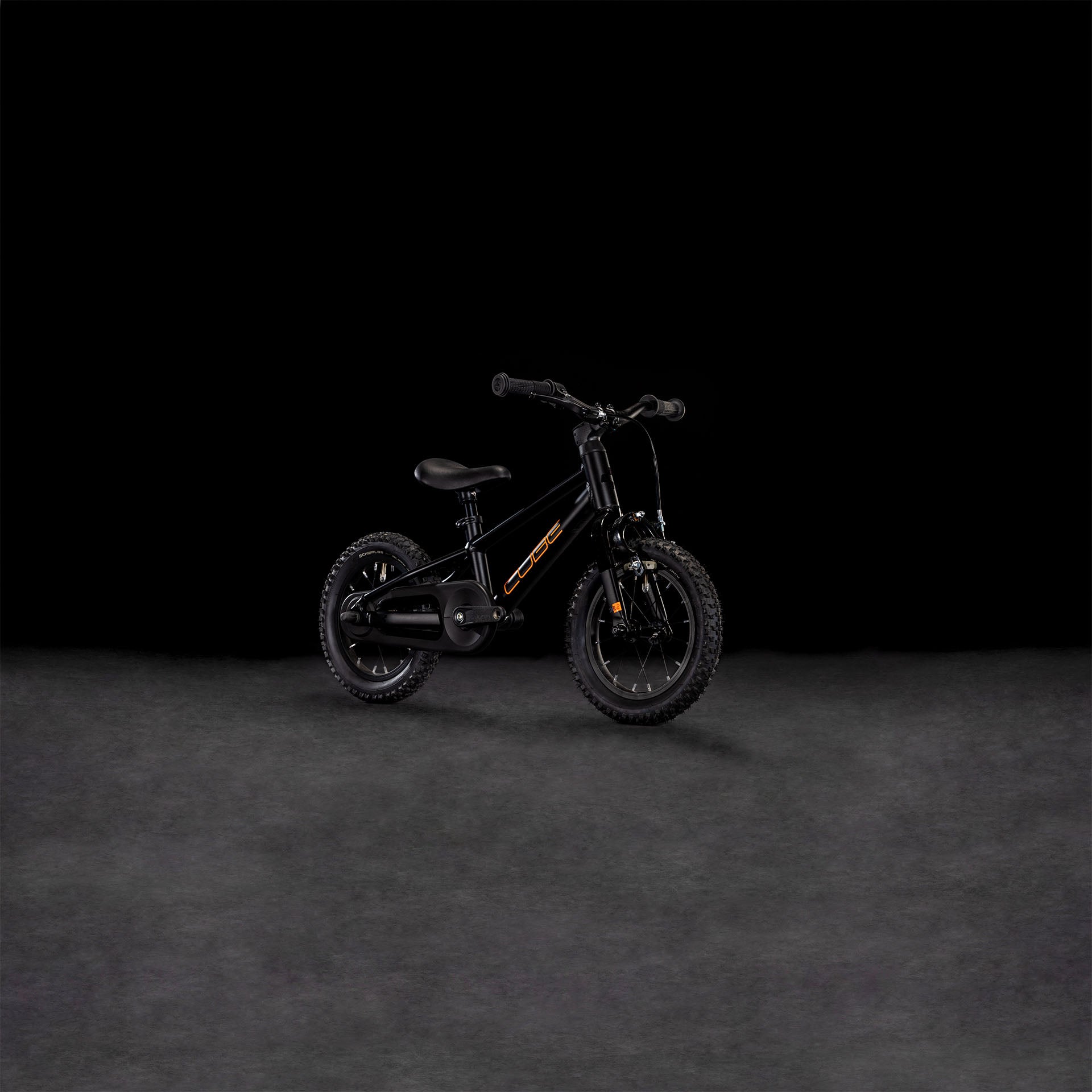Das CUBE Numove 120 RT black'n'orange 2025, ein kleines Kinderfahrrad mit Stützrädern, grobstolligen Reifen, niedrigem Rahmen und Felgenbremse für sicheres Anhalten, steht mittig vor einem dunklen Hintergrund.