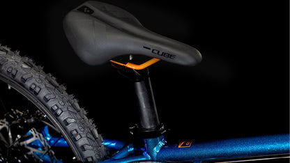 Nahaufnahme des schwarzen Sattels und der Sattelstütze des CUBE Acid 200 Disc Actionteam 2025 von CUBE. Zu sehen ist ein Teil eines dicken, grobstolligen Hinterreifens und ein metallicblauer Rahmen mit Shimano 7-fach-Schaltung, alles vor einem schwarzen Hintergrund.