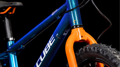 Nahaufnahme des CUBE Acid 200 Disc Actionteam 2025 mit blauem Rahmen, orangefarbener Vordergabel, Shimano 7-Gang-Schaltung, schwarzem Mountainbike-Reifen und sichtbarem CUBE-Logo auf schwarzem Hintergrund.