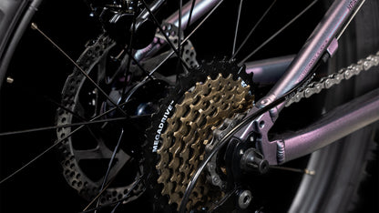 Nahaufnahme des Hinterrads des CUBE Acid 200 Disc galactic´n´cyan 2025. Shimano 7-Gang-Schaltung, Kassette, Kette, Speichen und hydraulische Scheibenbremsen sind zu sehen. Der metallische CUBE-Rahmen glänzt violett vor dunklem Hintergrund.