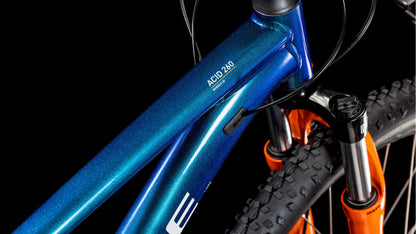Nahaufnahme des CUBE Acid 260 Disc actionteam 2025 Mountainbike-Rahmens in Blau mit schwarzen Zügen und einer orangefarbenen Federgabel mit Noppenreifen vor einem schwarzen Hintergrund.