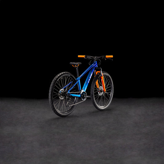Das CUBE Acid 260 Disc actionteam 2025 Mountainbike verfügt über einen Rahmen aus 6061er Aluminium, schwarze Reifen und einen orangefarbenen Lenker, der sich auf einer dunkel strukturierten Oberfläche vor einem schwarzen Hintergrund präsentiert.