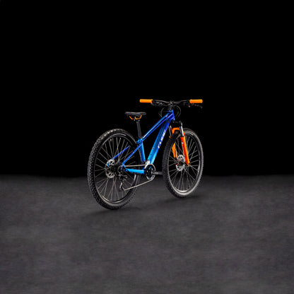 Das CUBE Acid 260 Disc actionteam 2025 Mountainbike verfügt über einen Rahmen aus 6061er Aluminium, schwarze Reifen und einen orangefarbenen Lenker, der sich auf einer dunkel strukturierten Oberfläche vor einem schwarzen Hintergrund präsentiert.