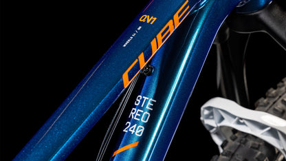 Nahaufnahme des metallisch blauen Cube Stereo 240 ONE actionteam 2025 Rahmens von CUBE, mit "CUBE" und "STEREO 240" in orange und weiß, mit einem Teil des Hinterrads und der Kabelbefestigung an diesem 24 Zoll Trailbike.
