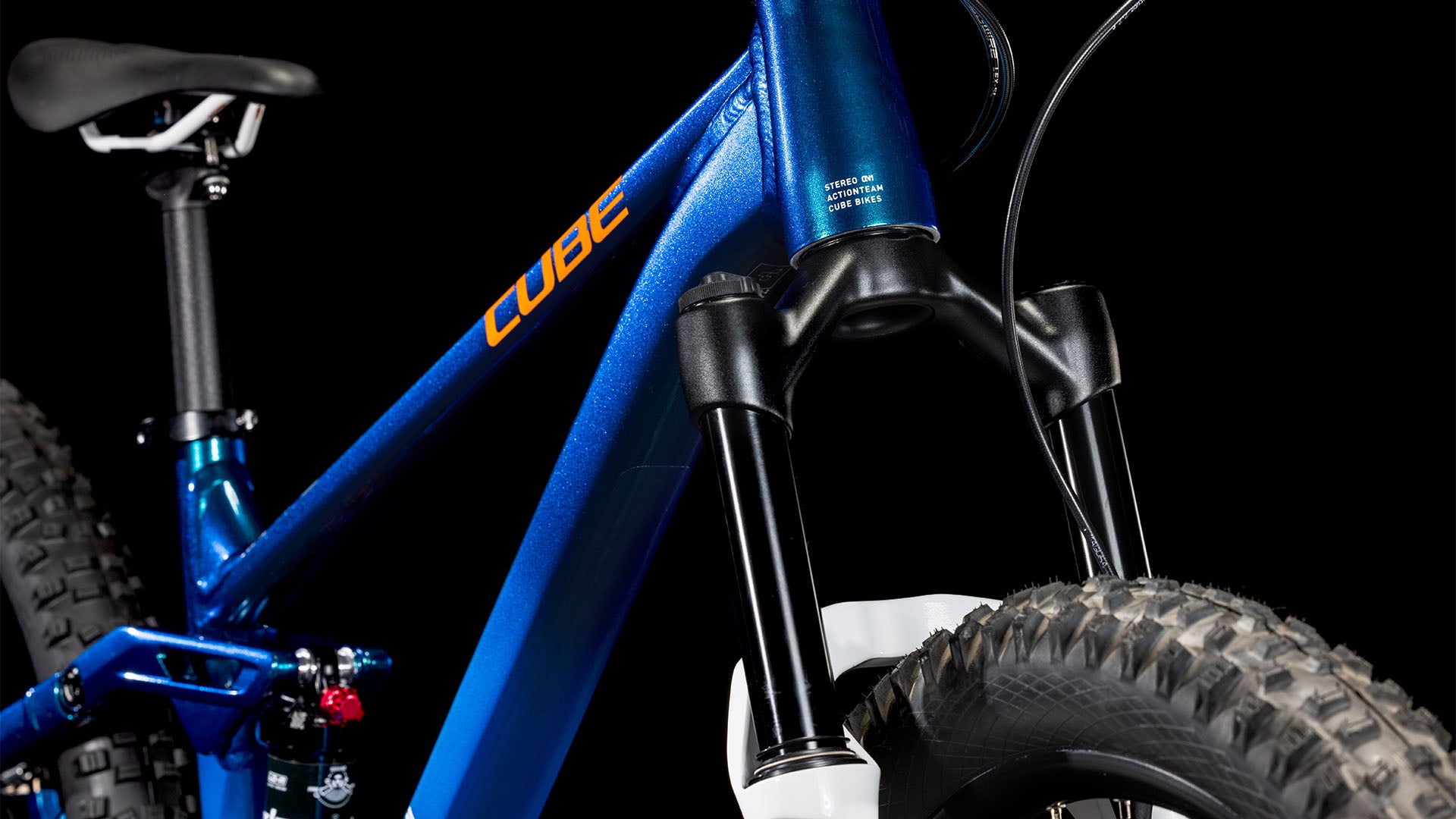 Nahaufnahme eines blauen CUBE Stereo 240 ONE actionteam 2025 Kinder-Mountainbikes, die die vordere Federgabel, den dicken Vorderreifen, den Teilrahmen und den Sattel vor einem schwarzen Hintergrund zeigt.